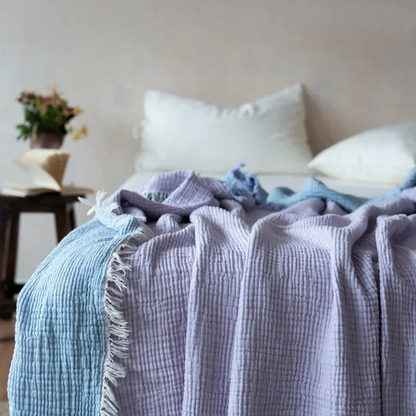 A lavender crinkled cotton throw blanket with a fringed edge spread over a bed with white pillows.