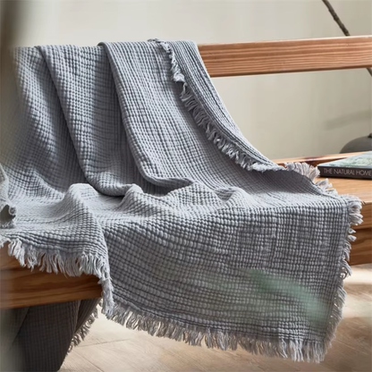 A gray crinkled cotton throw blanket with a fringed edge draped over a wooden bench.