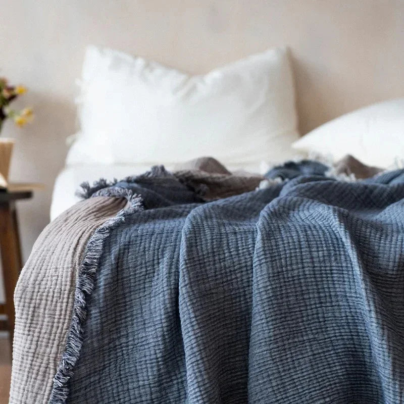 A slate blue-gray crinkled cotton throw blanket with a fringed edge spread over a bed with white pillows.
