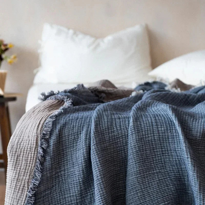 A slate blue-gray crinkled cotton throw blanket with a fringed edge spread over a bed with white pillows.