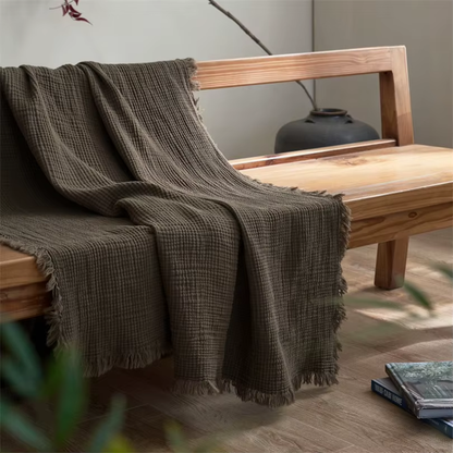 An olive crinkled cotton throw blanket with a fringed edge draped over a wooden bench.
