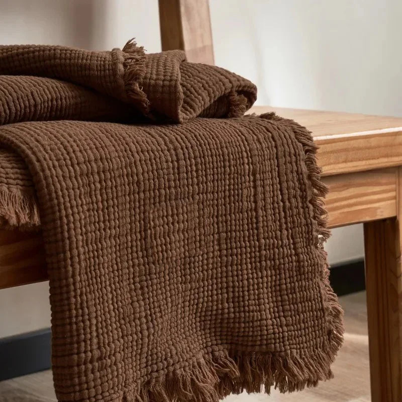 A brown crinkled cotton throw blanket with a fringed edge folded over a wooden bench.
