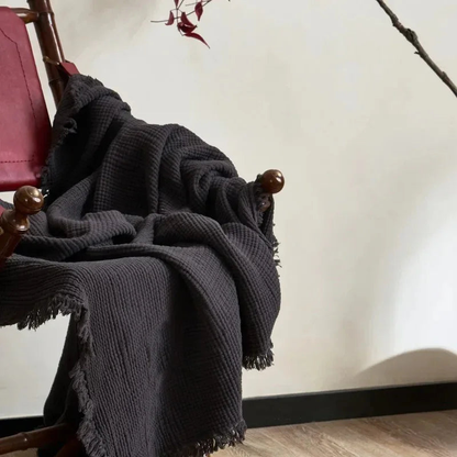 A charcoal crinkled cotton throw blanket with a fringed edge thrown over a chair.