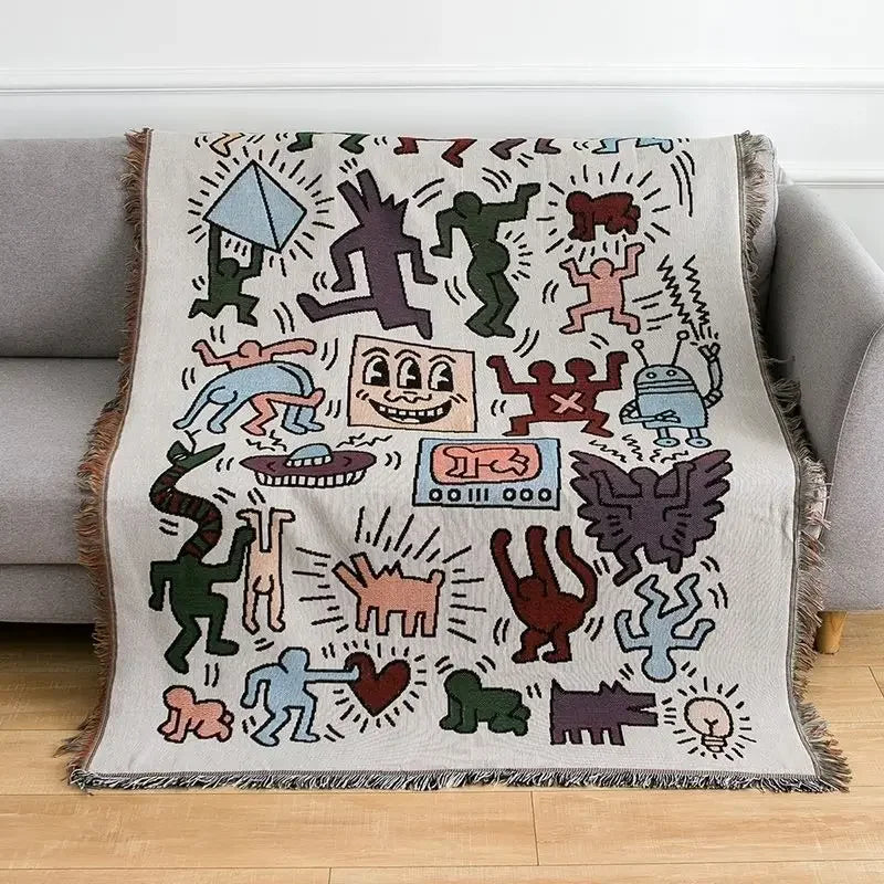 A woven throw blanket with black-outlined doodle figures in green, red, plum and blue hanging over the back and seat of a gray couch.