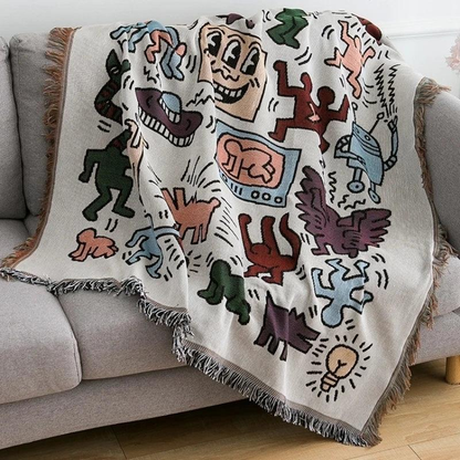 A fringed doodle throw blanket thrown at an angle across a gray couch, one corner falling toward a wood floor.