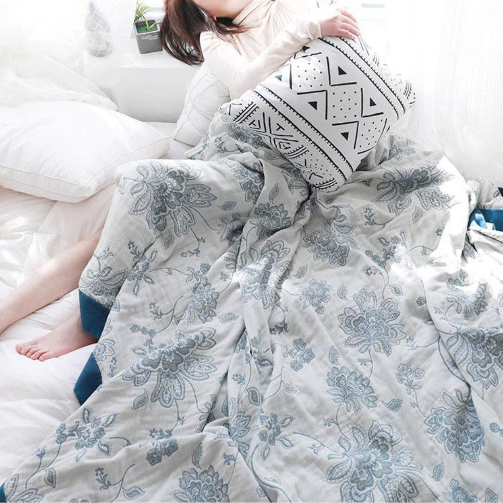 Woman sitting up in bed holding a cushion, the blue floral gauze blanket loose over her legs