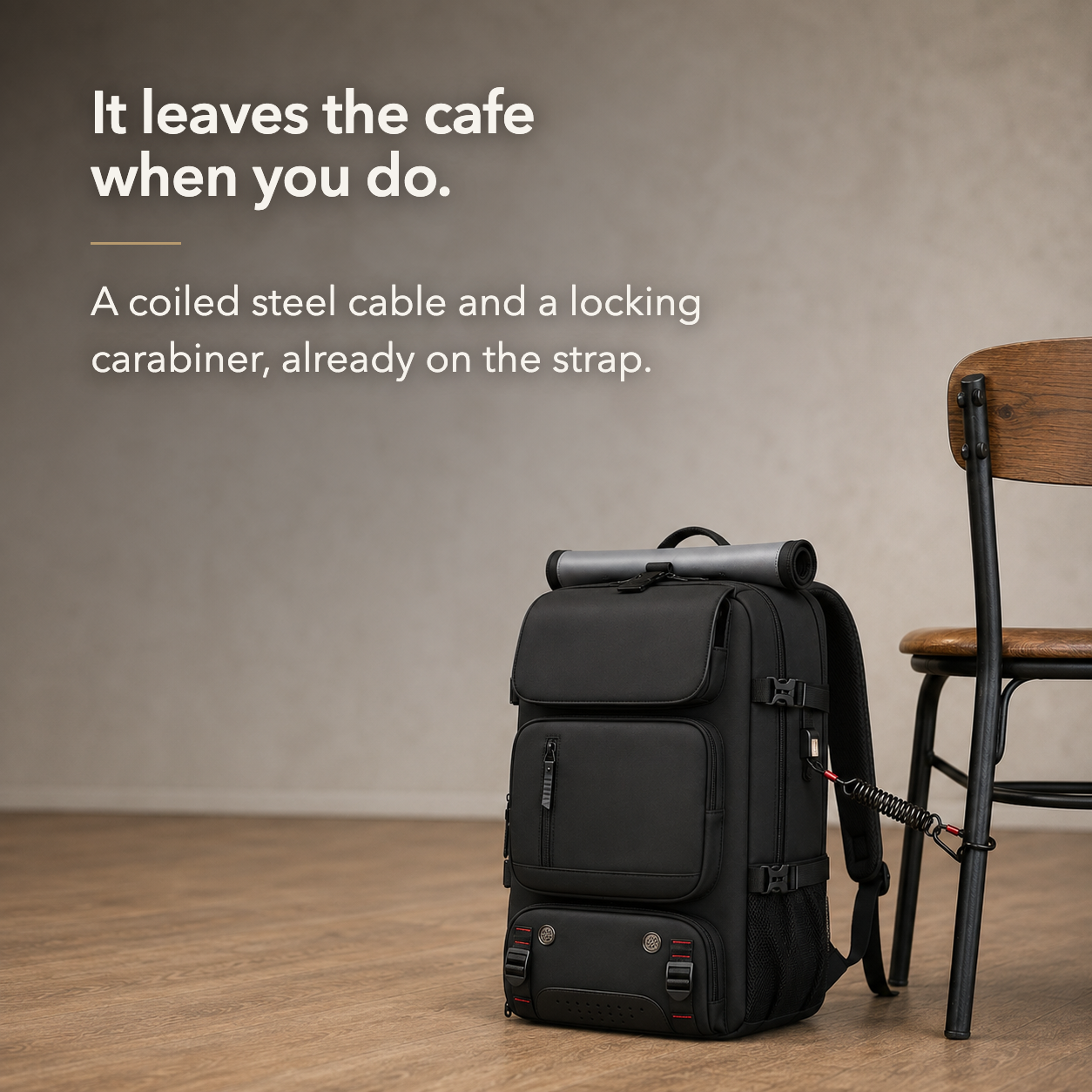 Black travel backpack on a cafe floor with its coiled steel cable and screw gate carabiner clipped around a chair leg