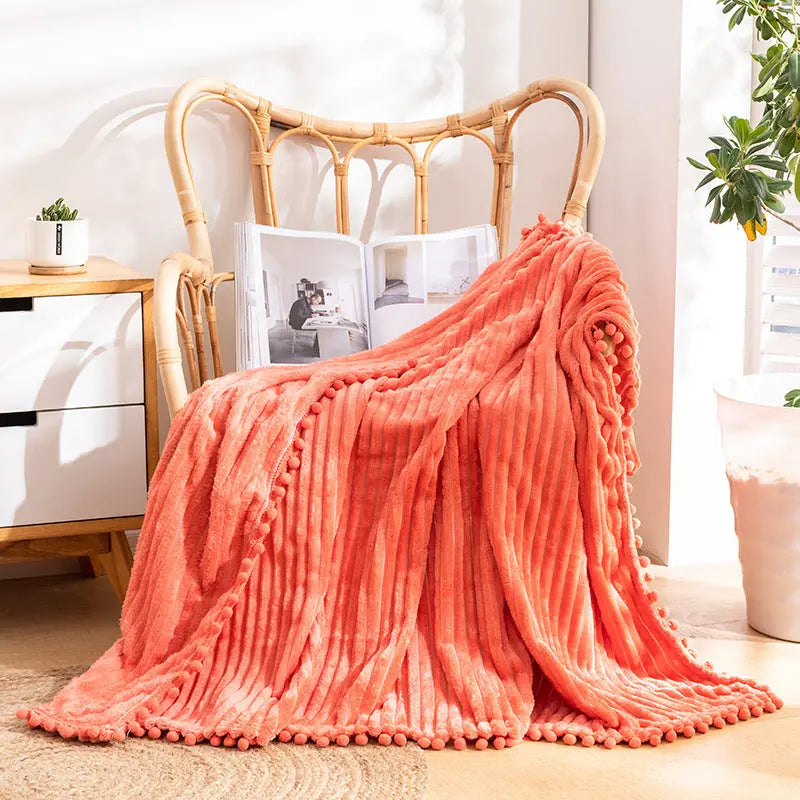 A coral flannel throw with raised stripes and coral pom-poms along its edge, over a rattan armchair