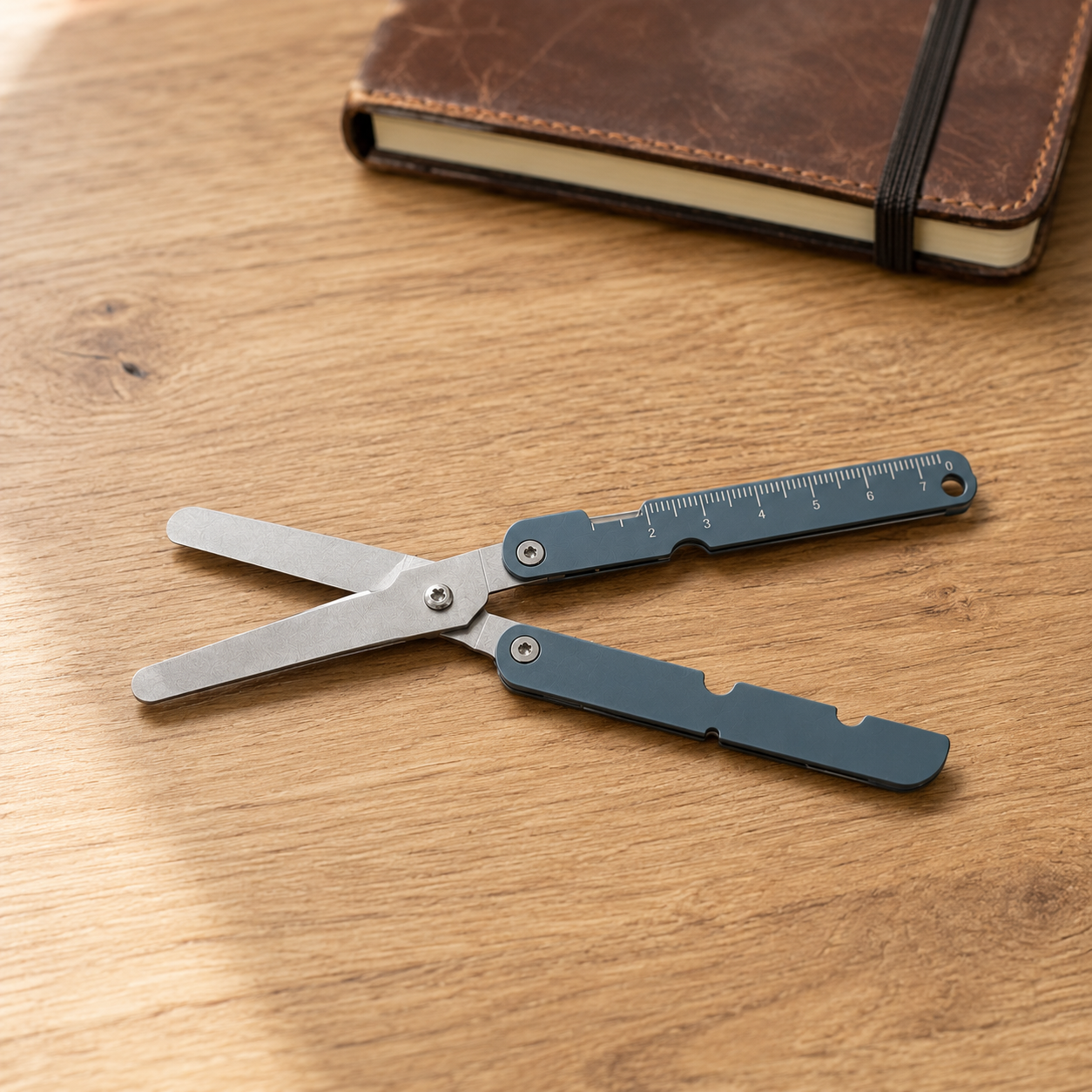 Gray blue folding pocket scissors open on a warm oak desk beside a notebook with an elastic closure.
