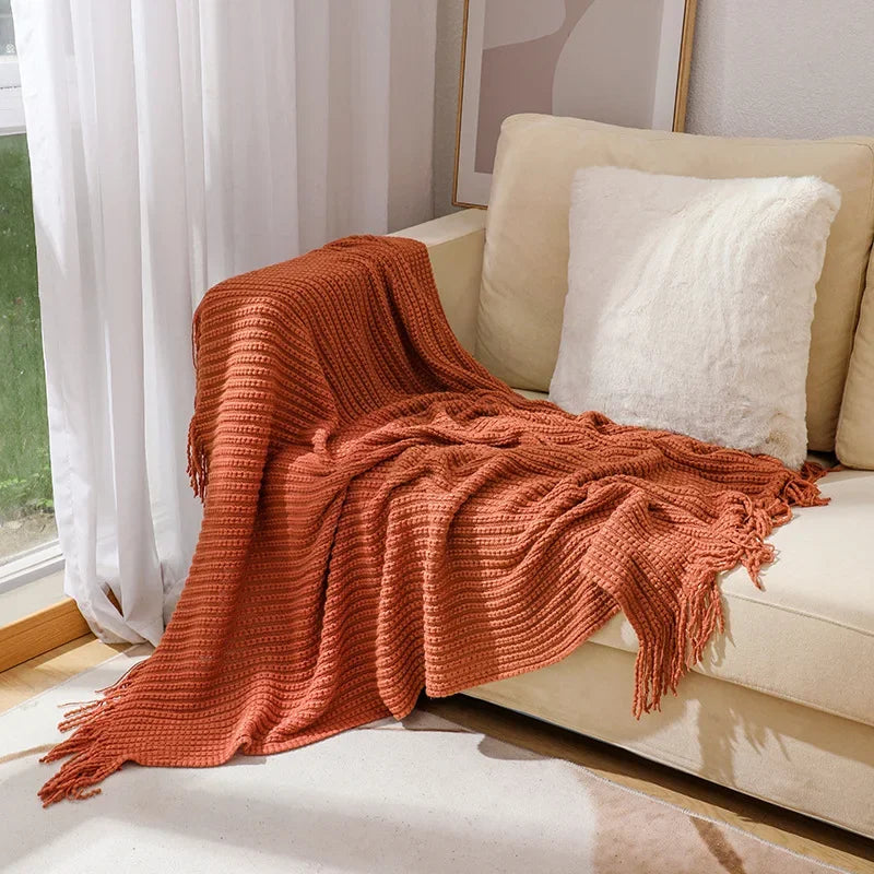 A rust bobble knit throw blanket with fringed ends spread over a sofa beside a sunny window.