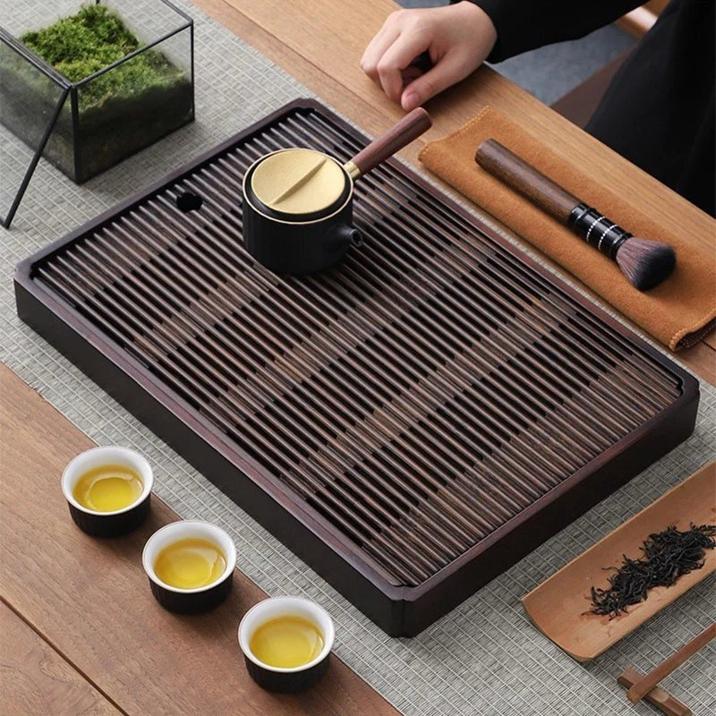 A side-handle teapot standing on the dark slats of the tea tray, with three cups of tea and a leaf scoop beside it.