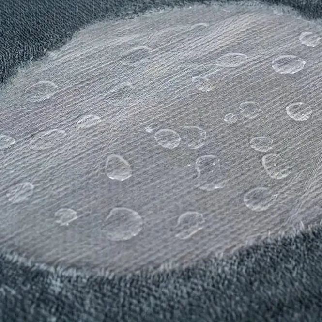 Close-up of a thin translucent film over dark gray pile, with water sitting on it in beads