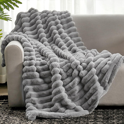 A gray faux fur throw blanket with long raised ribs, draped over the arm and seat of a pale sofa onto a patterned rug.