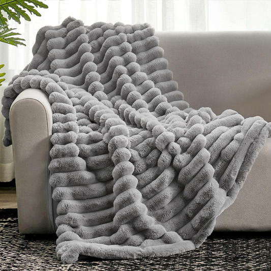 A gray faux fur throw blanket with long raised ribs, draped over the arm and seat of a pale sofa onto a patterned rug.