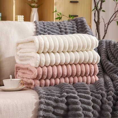 Folded cream and pink ribbed faux fur throws stacked on a sofa, with a charcoal one spread out behind them.
