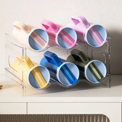 A clear two-tier bottle rack holding six large tumblers with handles on their sides, standing on a white sideboard.