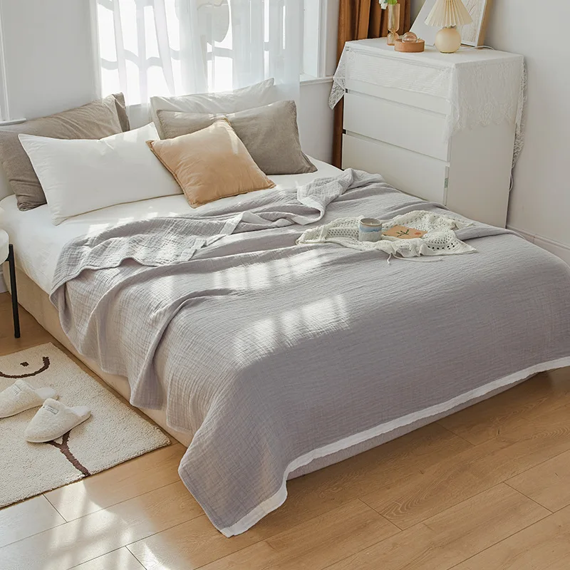 Gray muslin cotton blanket spread over a double bed in a sunlit bedroom, its white edge band running along the hem
