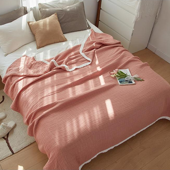 Coral muslin blanket spread over a bed from the foot, white band along the edges, a book and tulips on top
