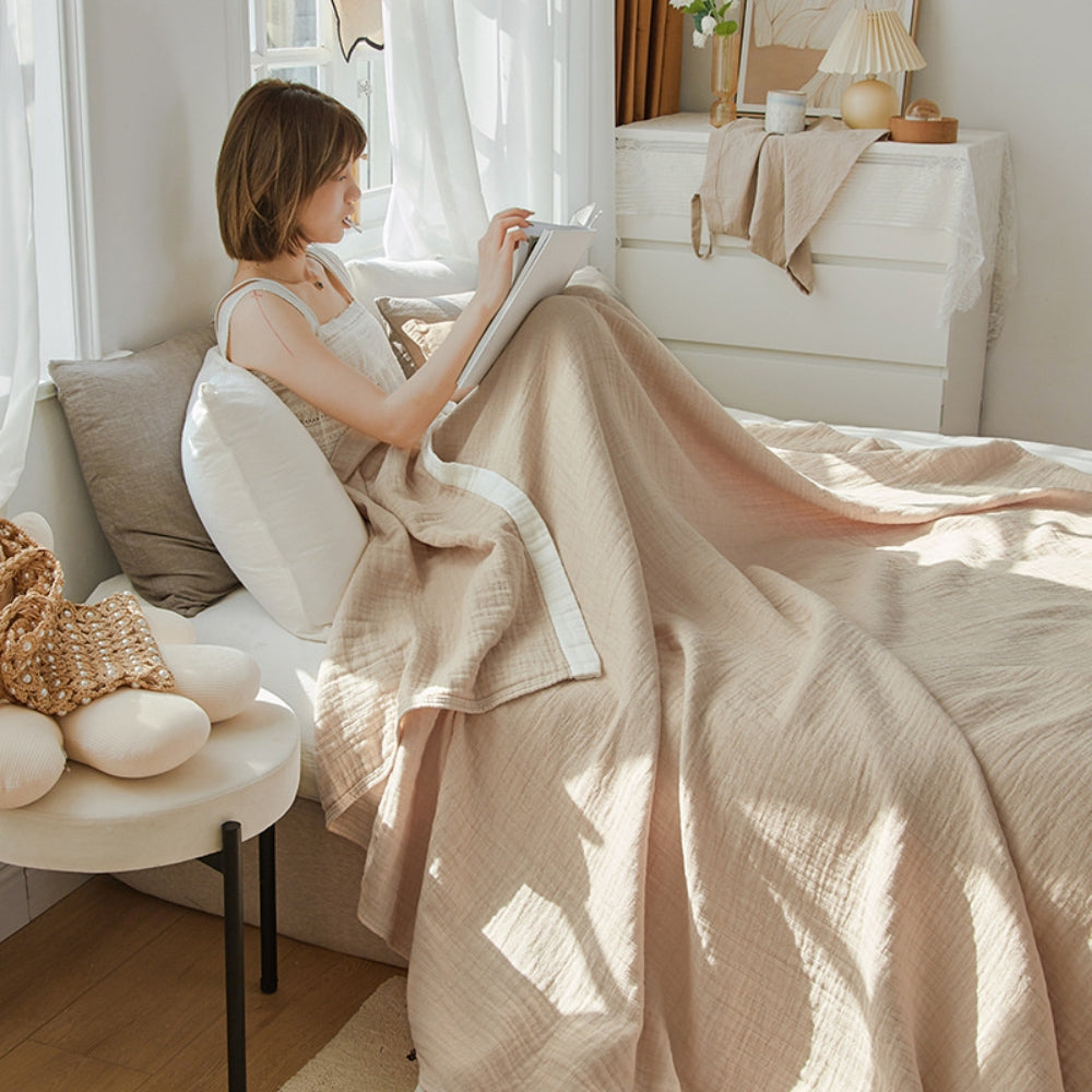 Woman reading in bed under a beige muslin cotton blanket that spills over the side of the bed