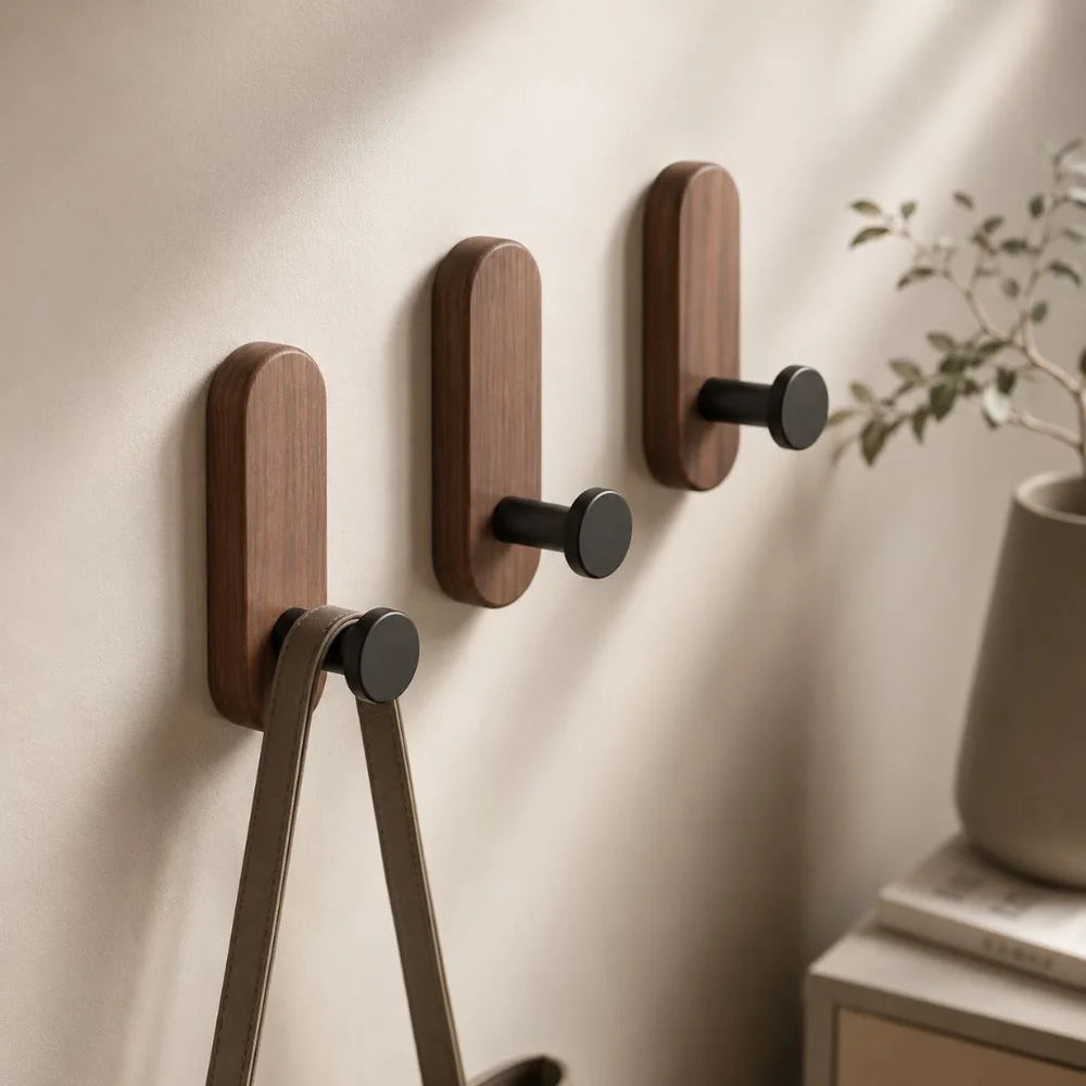Three walnut wall hooks with black pegs on a warm white wall, a leather tote strap hanging from the lowest