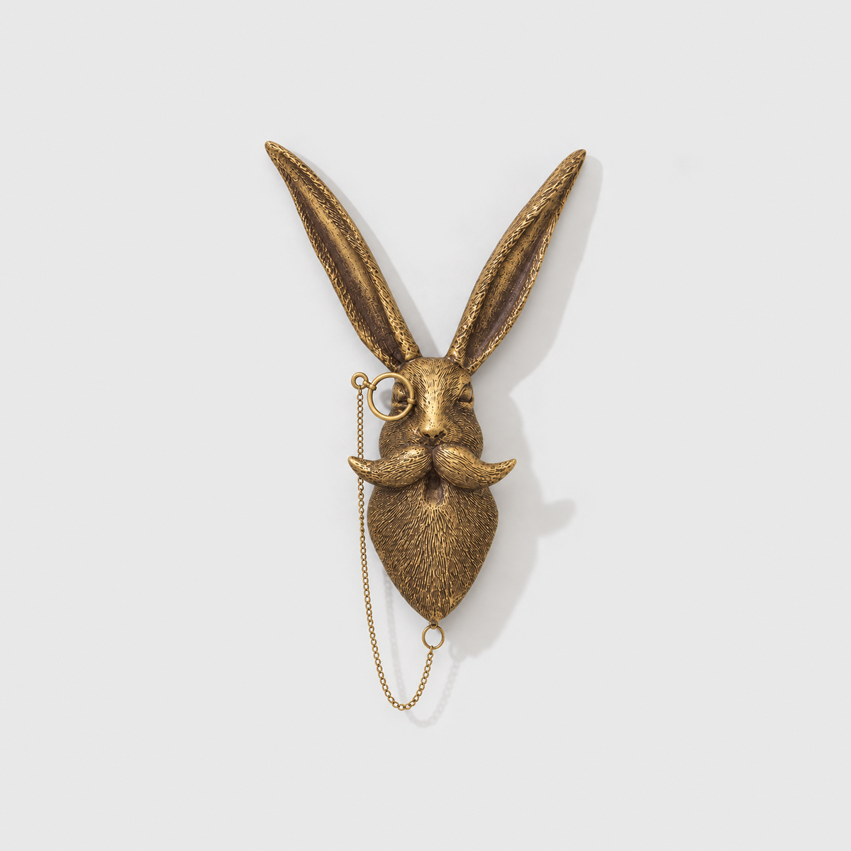 Rabbit head wall sculpture in antique gold-tone resin, with a monocle on a chain, on a plain light gray wall