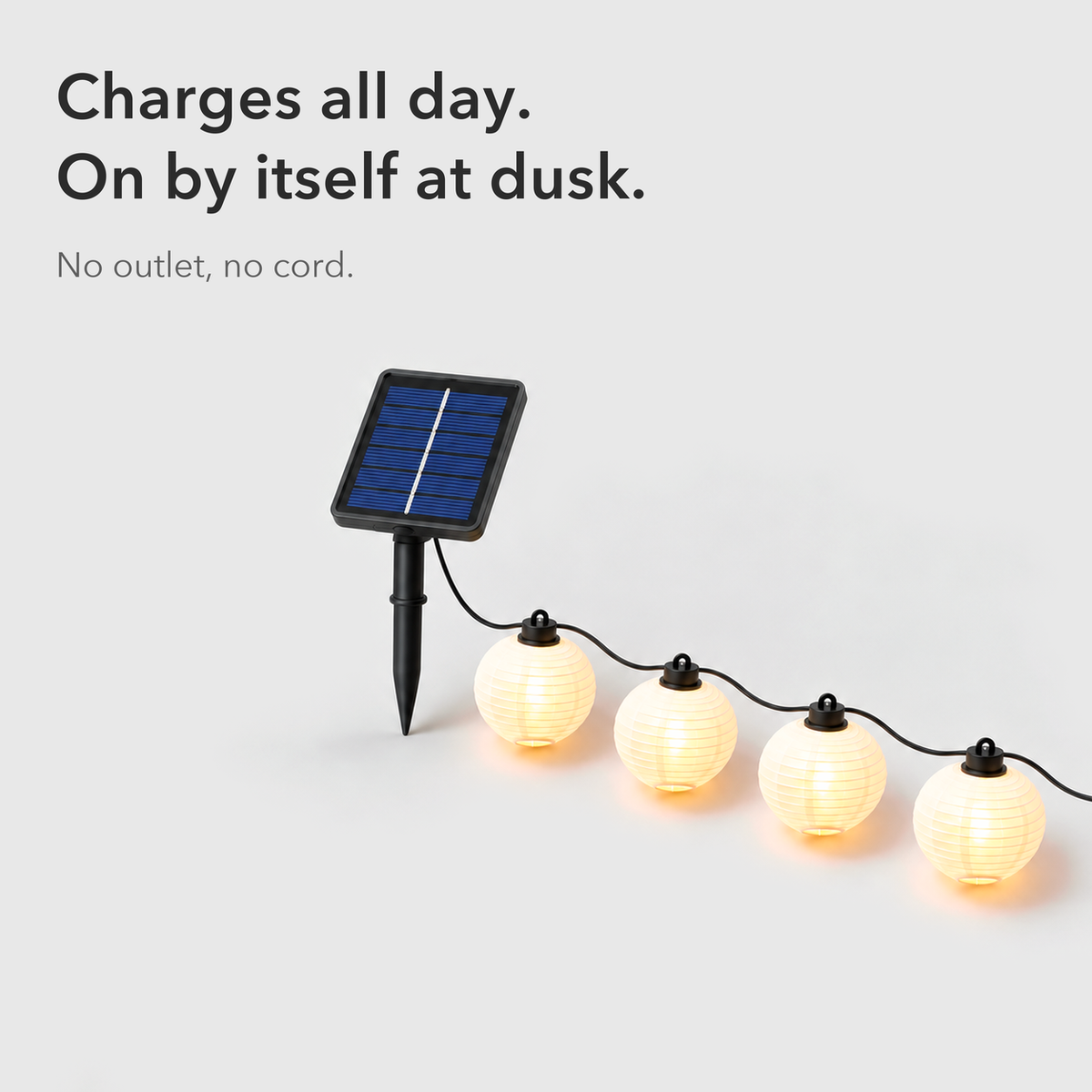 Text card reading Charges all day, on by itself at dusk, beside the square solar panel on its stake and the first white lanterns of the string