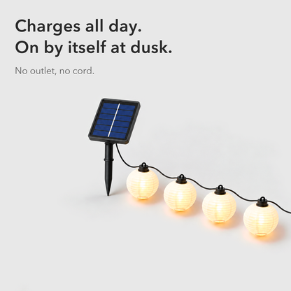 Text card reading Charges all day, on by itself at dusk, beside the square solar panel on its stake and the first white lanterns of the string