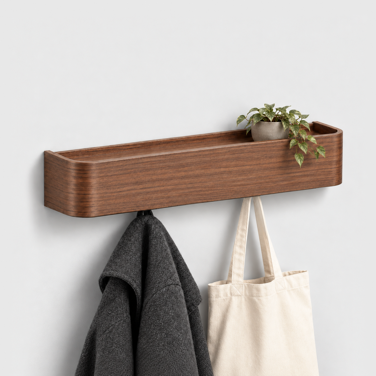 Walnut wall coat rack with shelf, a small trailing plant in a gray pot on top and a charcoal coat and canvas tote bag hanging from the hooks underneath