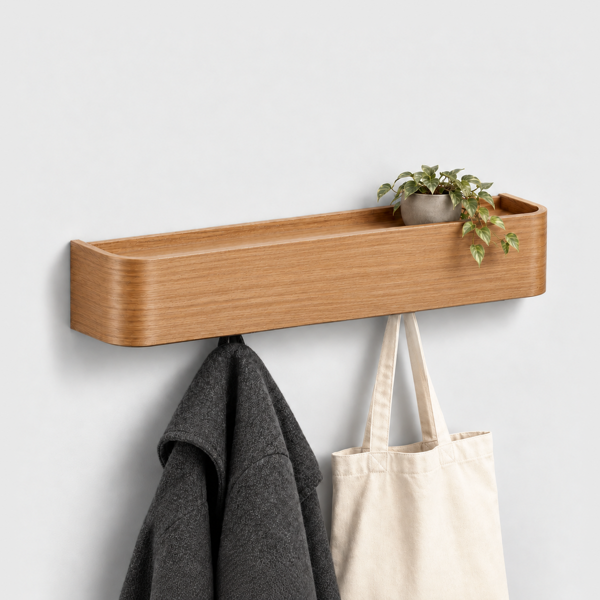 Natural wood wall coat rack with shelf, a small trailing plant in a gray pot on top and a charcoal coat and canvas tote bag hanging from the hooks underneath