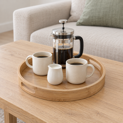 Round bamboo serving tray holding a French press, two mugs and a small milk jug on an oak coffee table