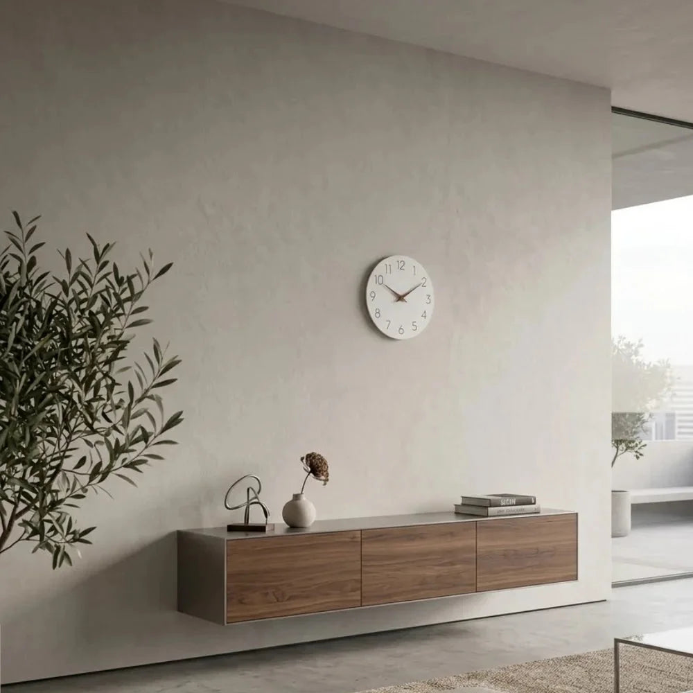 A small white wall clock above a floating walnut cabinet on a large plaster wall