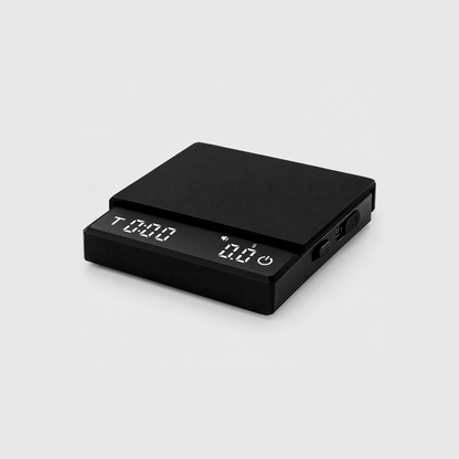 Black coffee scale with its silicone mat on top and the display lit showing the timer and 0.0 grams, on a light gray studio background