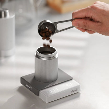 Coffee beans poured from a scoop into an aluminum dosing cup on the white coffee scale
