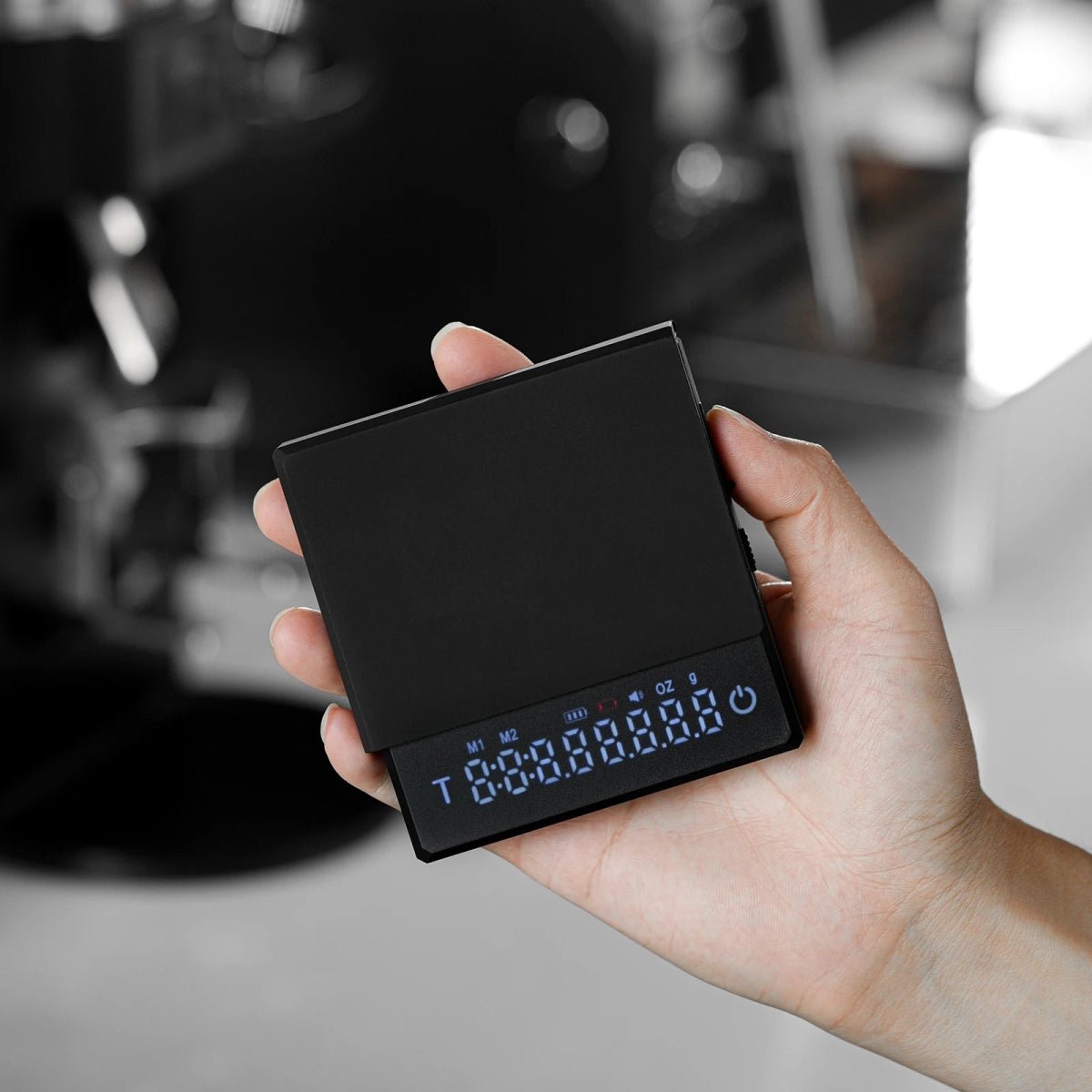 Black coffee scale held in one hand with every display segment lit, including the timer, mode and battery icons