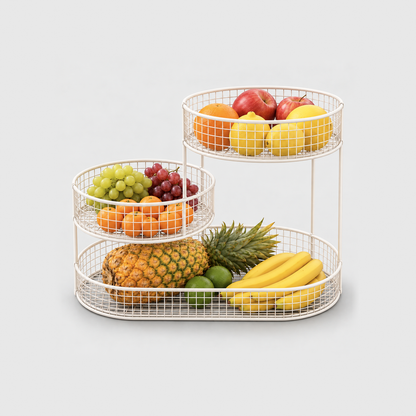 White 3 tier fruit basket stand filled with pineapple, bananas, grapes, oranges, lemons and apples, on a plain light gray background