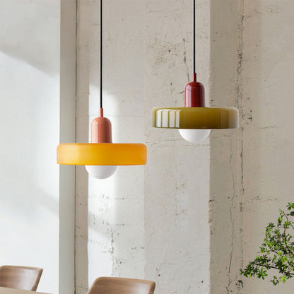 Two Bauhaus pendant lights, an amber glass shade on a pink body and an olive shade on a burgundy body, beside a concrete wall