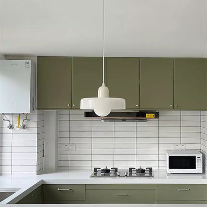 White glass pendant light with a beige body hanging over a kitchen with green cabinets and white tiles