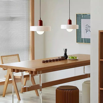 Two white glass pendants with burgundy bodies over a long wooden dining table by a window