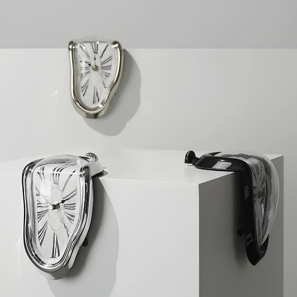 Three melting clocks in silver and black, two draped over the edge of a white block and one on the wall above