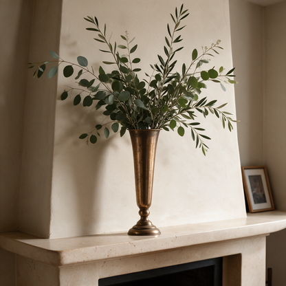Tall antique gold trumpet vase with eucalyptus branches on a plaster fireplace mantel