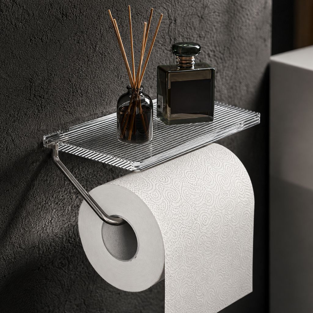 Silver toilet paper holder with a clear fluted acrylic shelf on a dark textured wall, with a diffuser and a dark bottle on the shelf