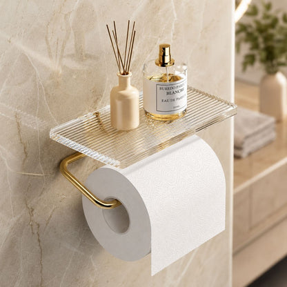 Gold toilet paper holder with a diffuser and a perfume bottle on its clear fluted shelf, on a cream marble wall