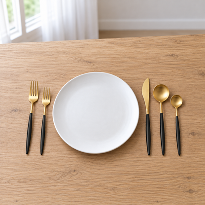 Black-handled gold flatware laid beside a white dinner plate on an oak table in daylight