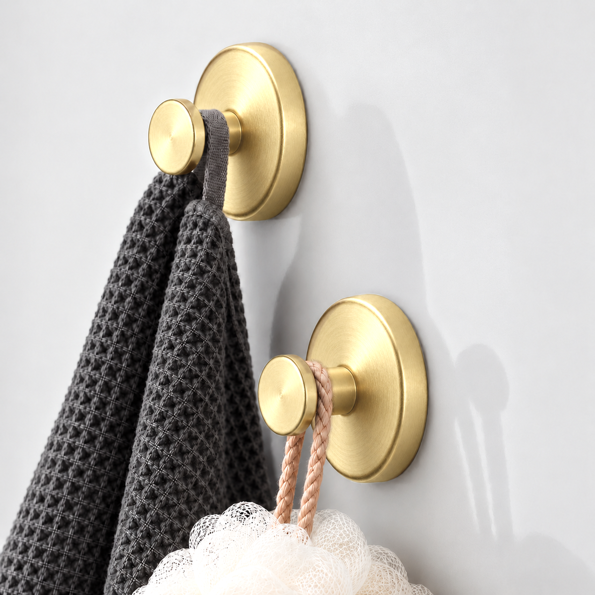Two brushed gold suction hooks seen from the side, a charcoal waffle cloth on the upper one and a white loofah on its cord on the lower one