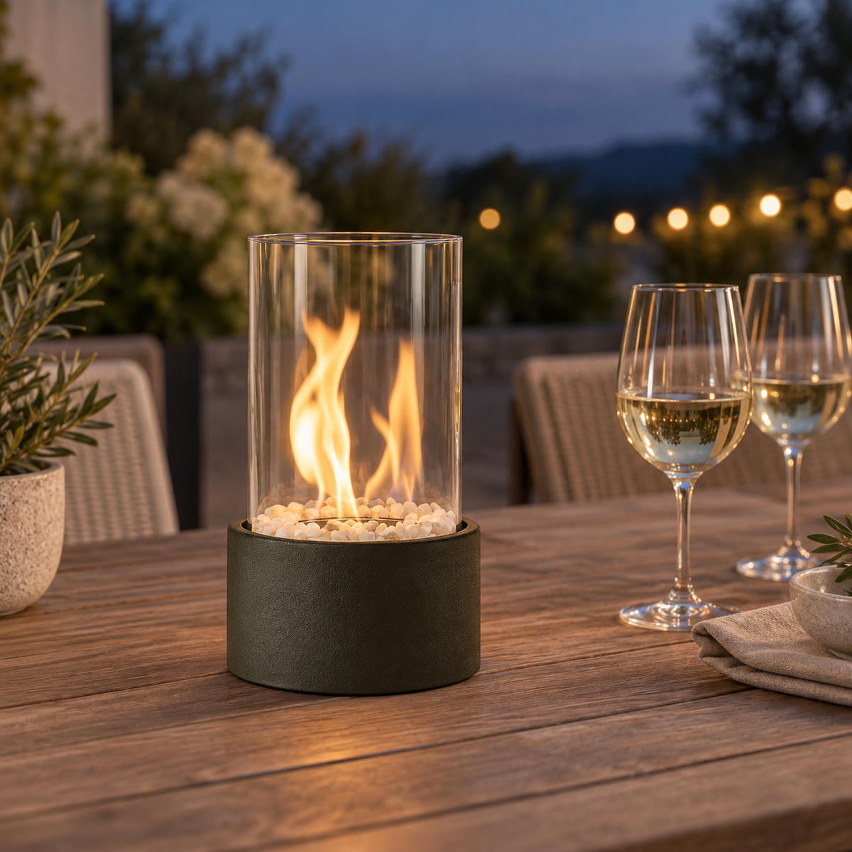 Black glass cylinder fire pit burning on a teak outdoor dining table at dusk beside two glasses of white wine