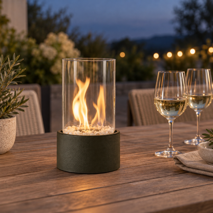 Black glass cylinder fire pit burning on a teak outdoor dining table at dusk beside two glasses of white wine