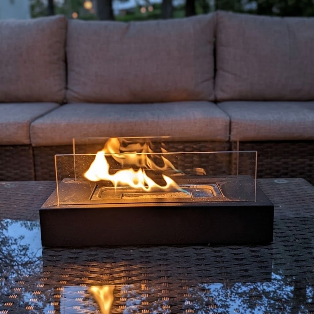 Rectangle tabletop fire pit burning between two glass panels on a glass-topped wicker table in front of an outdoor sofa at dusk