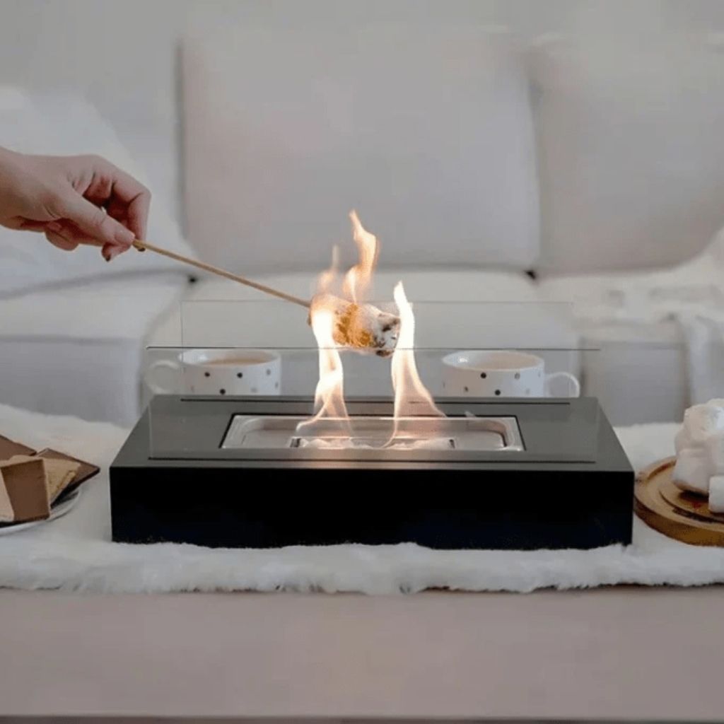 A hand toasting a marshmallow over the lit tabletop fire pit on a white rug, with two mugs and s'mores beside it
