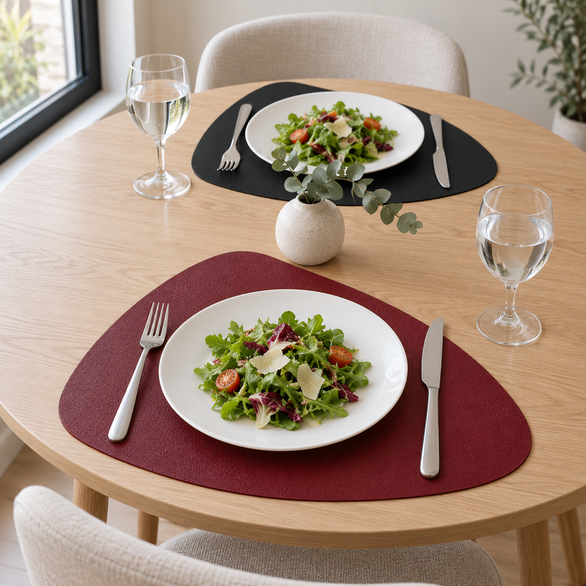 Two Burgundy and Black placemats on a round light wood table in daylight, one burgundy side up and one black