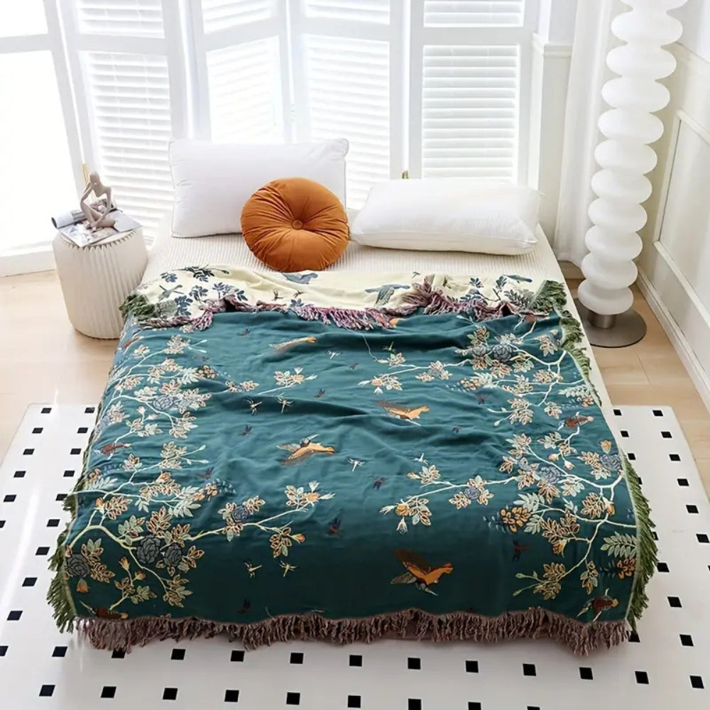 Teal bird and branch cotton blanket spread over a bed, its pale cream reverse folded back at the pillows and a fringe on every edge